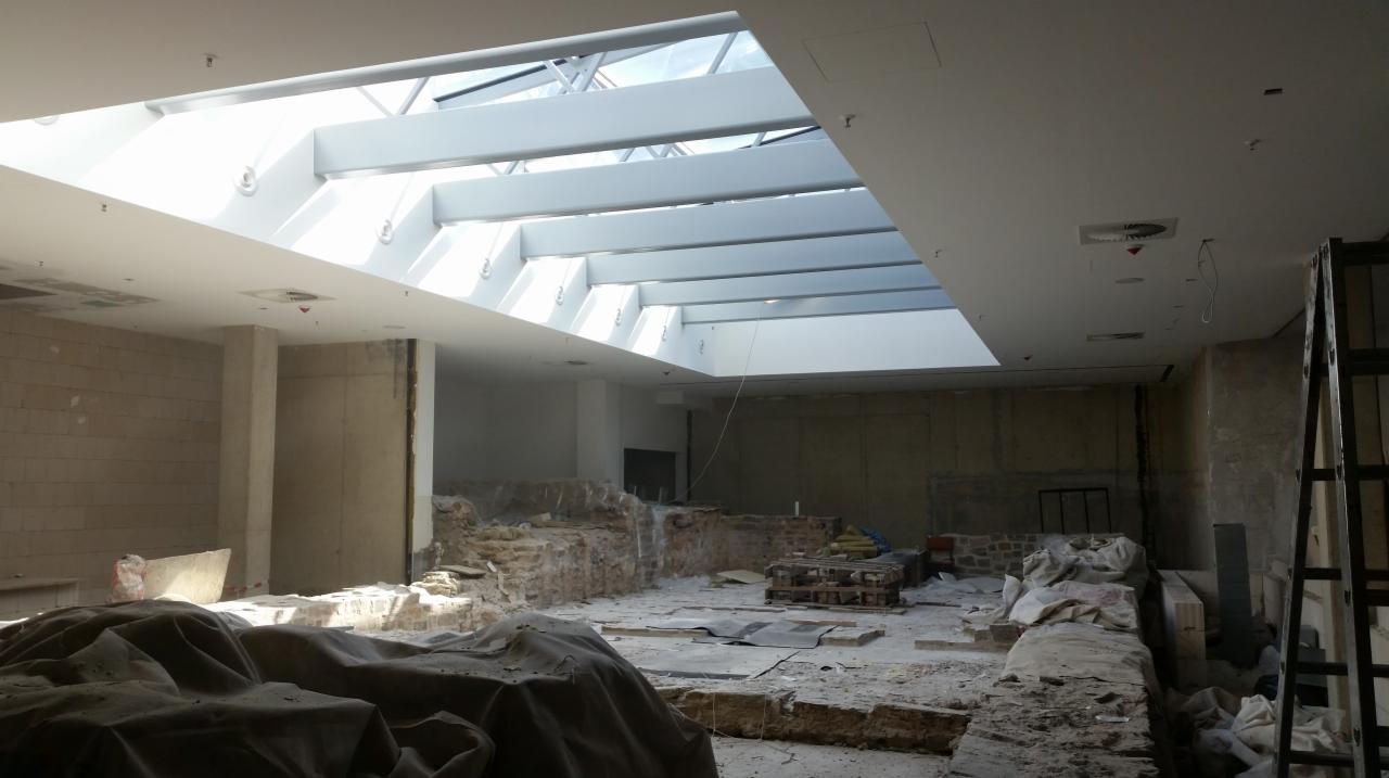 Wnętrze w trakcie remontu z odsłoniętymi fragmentami muru, przykryte plandekami, z widocznym przeszklonym sufitem doświetlającym przestrzeń.