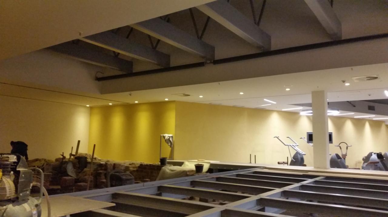 Wnętrze komercyjne w trakcie prac wykończeniowych; widoczne odsłonięte cegły, metalowa konstrukcja pod podłogą, żółte ściany i sprzęt do ćwiczeń w tle.