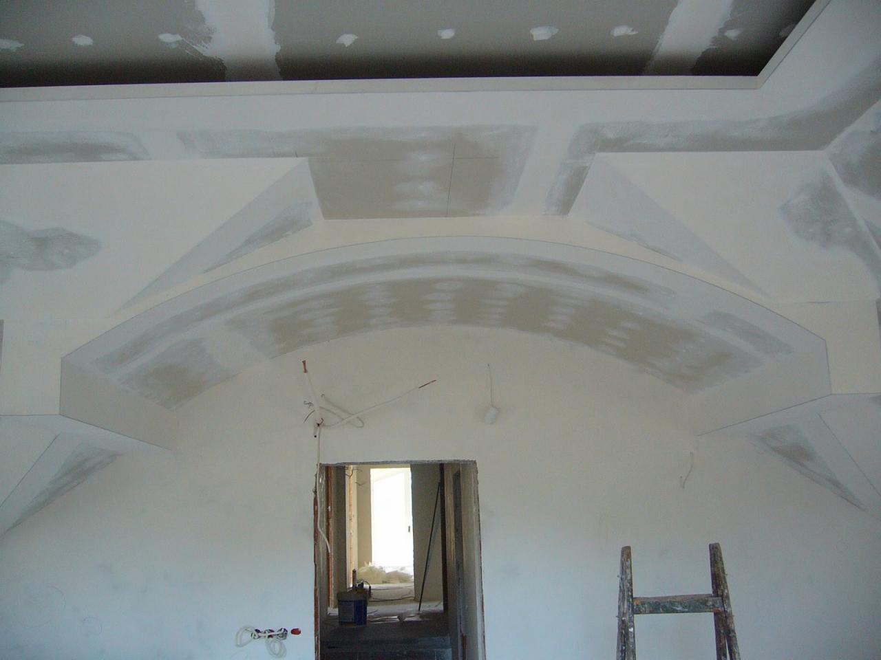 Wnętrze w trakcie wykańczania: widoczna konstrukcja łukowa z płyt na ścianie, częściowo pokryta masą wyrównującą, sufit z elementami dekoracyjnymi, przewody elektryczne wystające ze ściany, drabina...