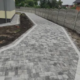 Długa, szara ścieżka z kostki brukowej prowadząca wzdłuż ogrodzenia panelowego, z obrzeżami z jasnego kruszywa, świeżo obsypana ziemią po bokach, widok z perspektywy.