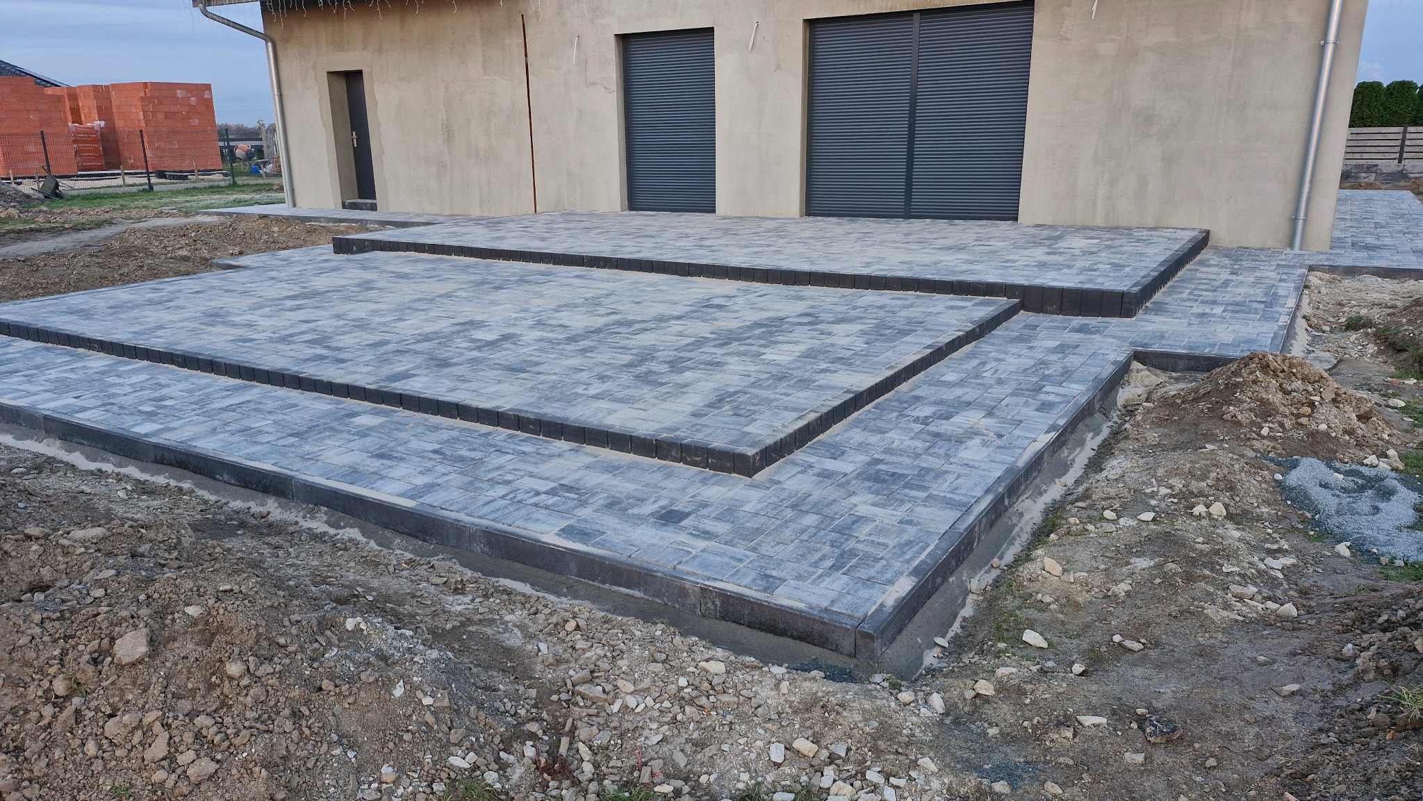 Nowoczesny taras z szarej kostki brukowej ułożonej schodkowo, z ciemnymi obrzeżami, na tle budynku z szarymi roletami. Widoczne ślady prac ziemnych.