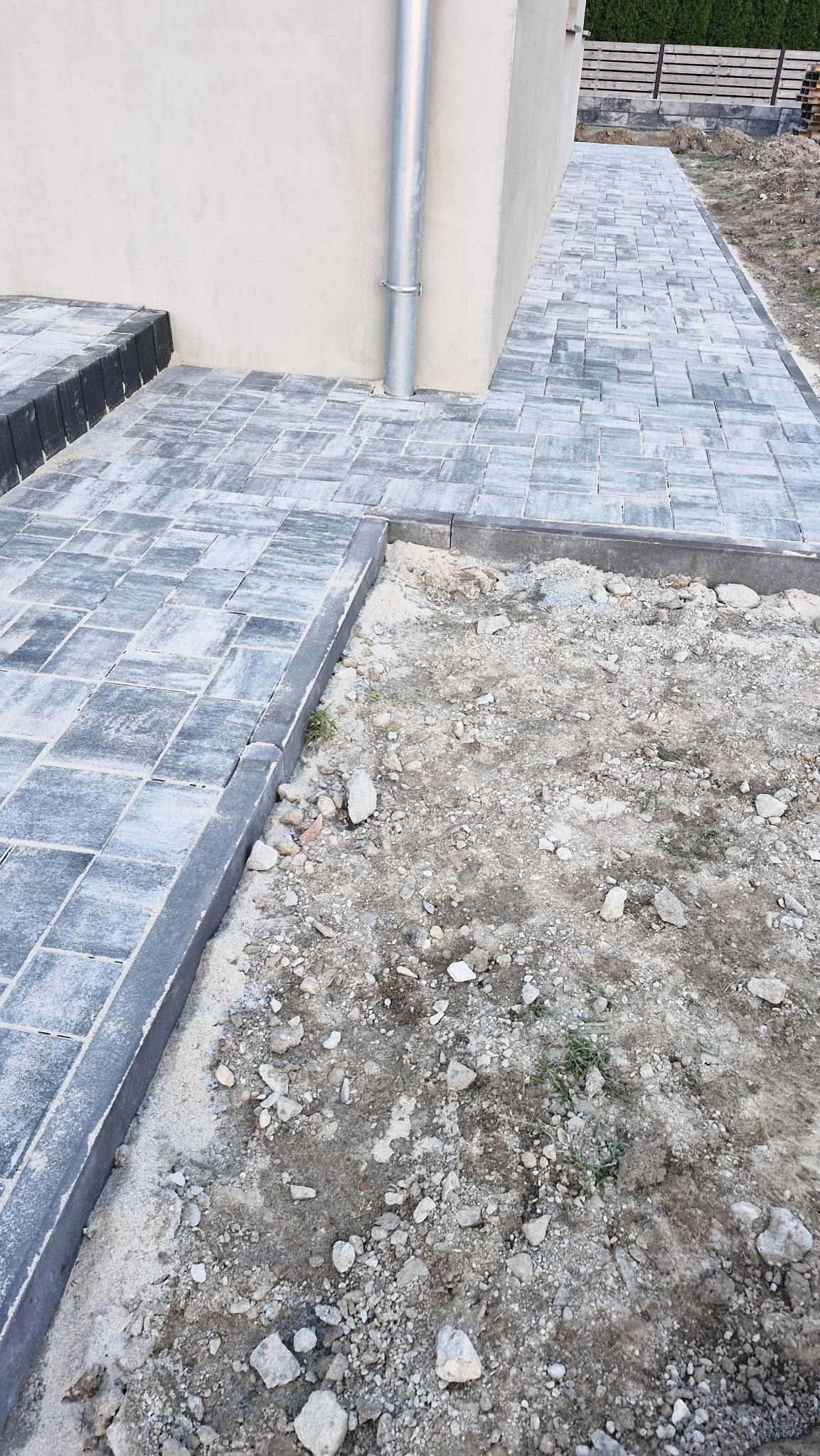 Ułożona szara kostka brukowa przy ścianie budynku z rynną, obok fragment terenu przygotowanego pod dalsze prace brukarskie. Widok z góry, perspektywa.