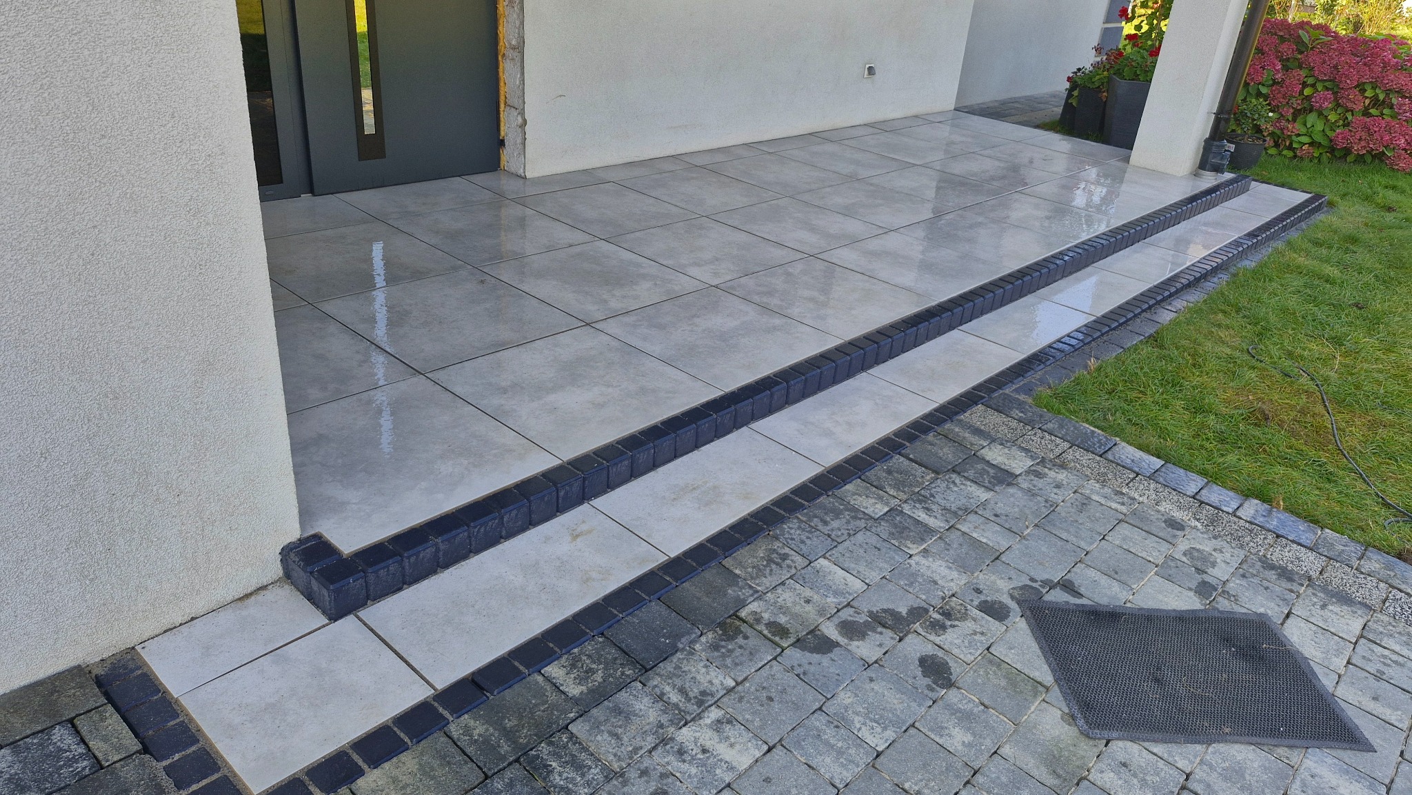 Nowoczesne schody wejściowe wyłożone jasnoszarą płytką, obrzeża z ciemnej kostki. Na dole kostka brukowa w odcieniach szarości, trawnik i fragment roślinności.