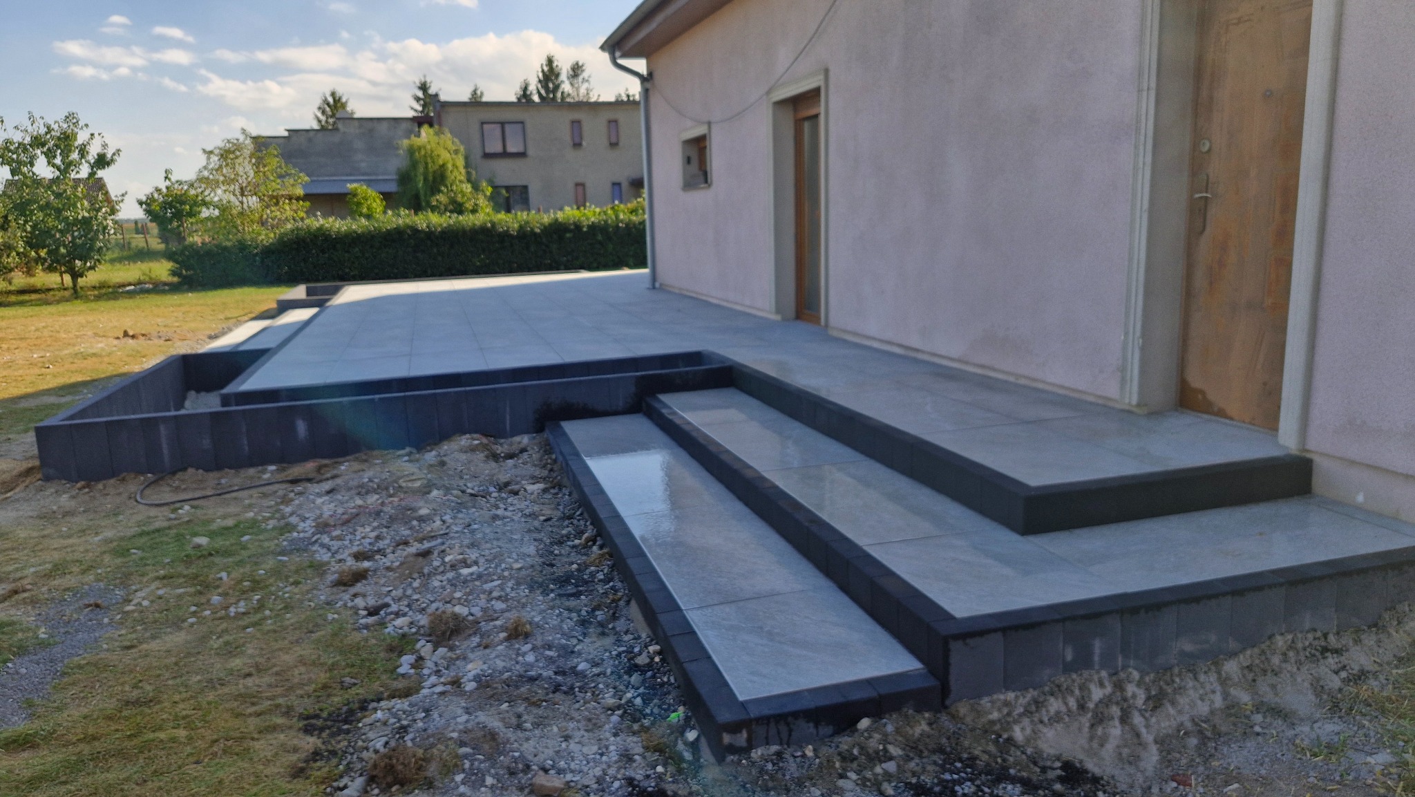 Nowoczesny taras z szarych płyt, z trzema schodami prowadzącymi do drzwi domu. Widoczne obrzeża z ciemnej kostki i fragment trawnika. Słoneczny dzień.