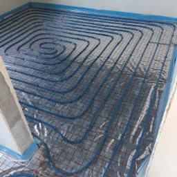 Niebieskie rury ogrzewania podłogowego ułożone spiralnie na folii z czerwoną kratką, widoczne fragmenty ścian w surowym stanie.