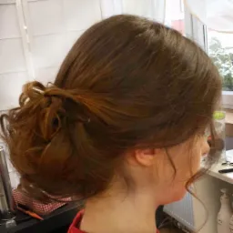 Upięcie włosów w stylu messy bun, brązowe włosy z delikatnymi refleksami, widok z profilu, w tle widać okno i regał z kosmetykami.