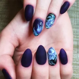 Dłoń z ciemnofioletowymi, matowymi paznokciami w kształcie migdała, z czego dwa zdobione ręcznie malowanymi niebieskimi kwiatami na czarnym tle i jeden z białymi kropkami u nasady.