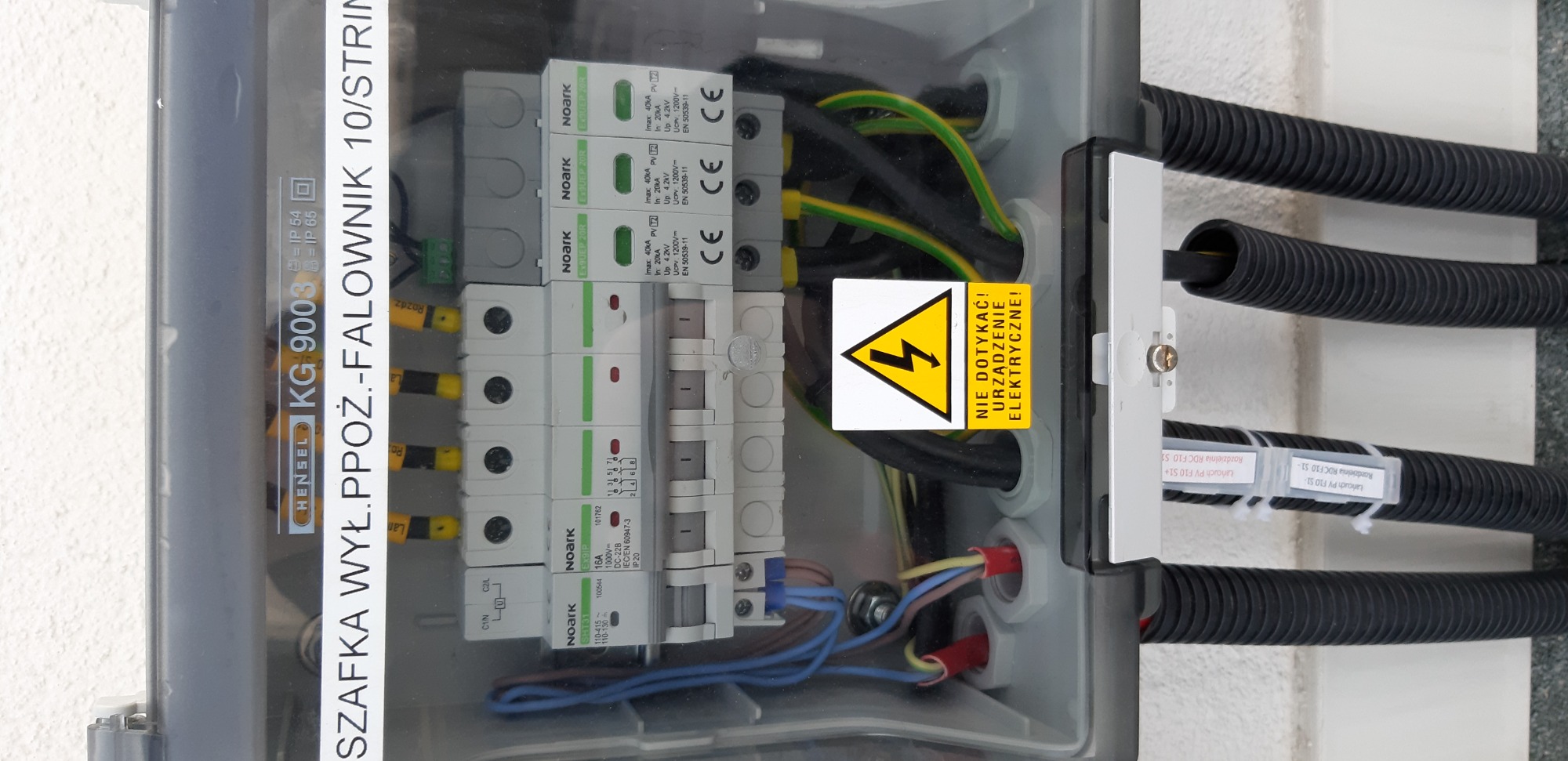 Otwarta skrzynka elektryczna z zabezpieczeniami przepięciowymi Noark, okablowaniem i żółtą naklejką ostrzegawczą 'Nie dotykać! Urządzenie elektryczne!', przytwierdzona do ściany, widoczne czarne...