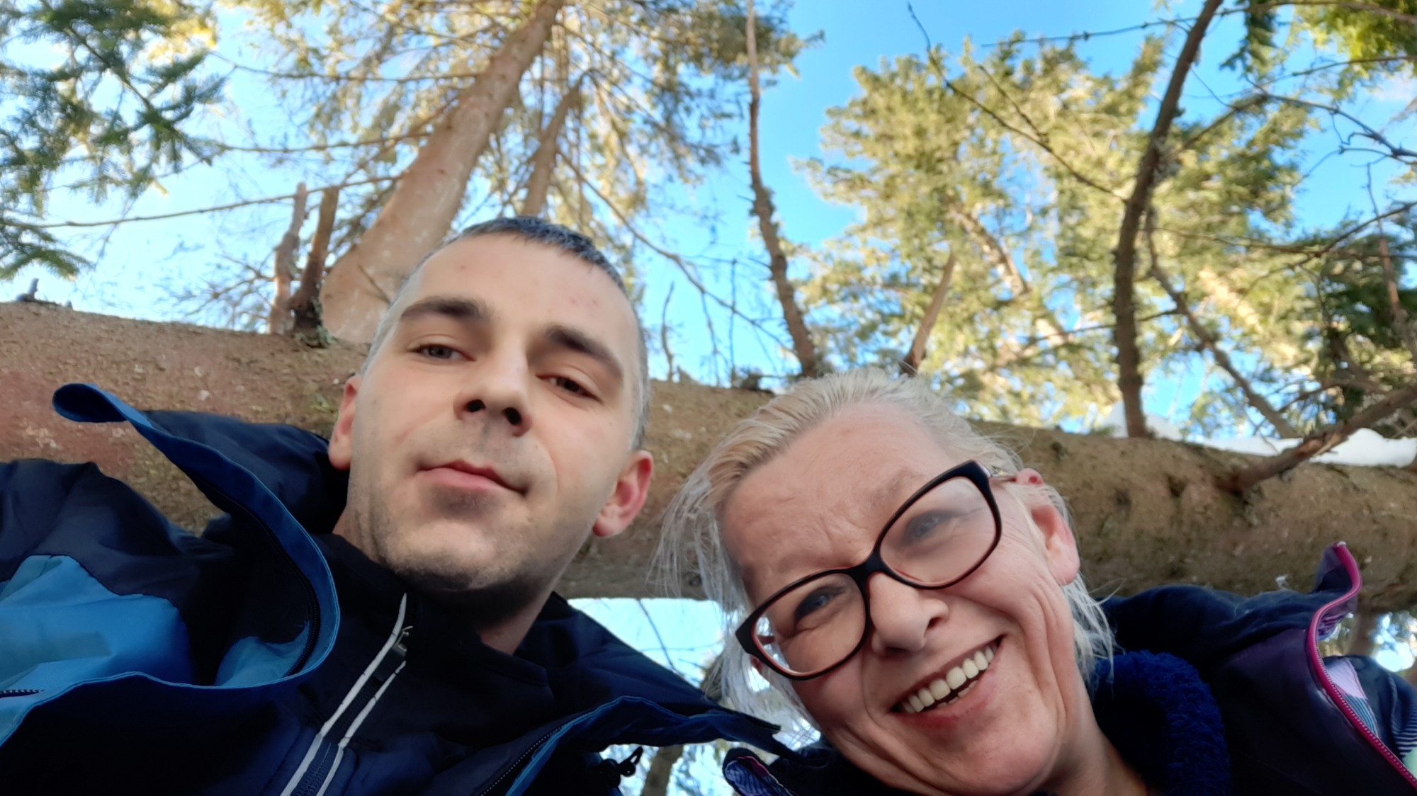 Selfie dwóch osób pod drzewem iglastym z fragmentami śniegu w tle, z widokiem na błękitne niebo