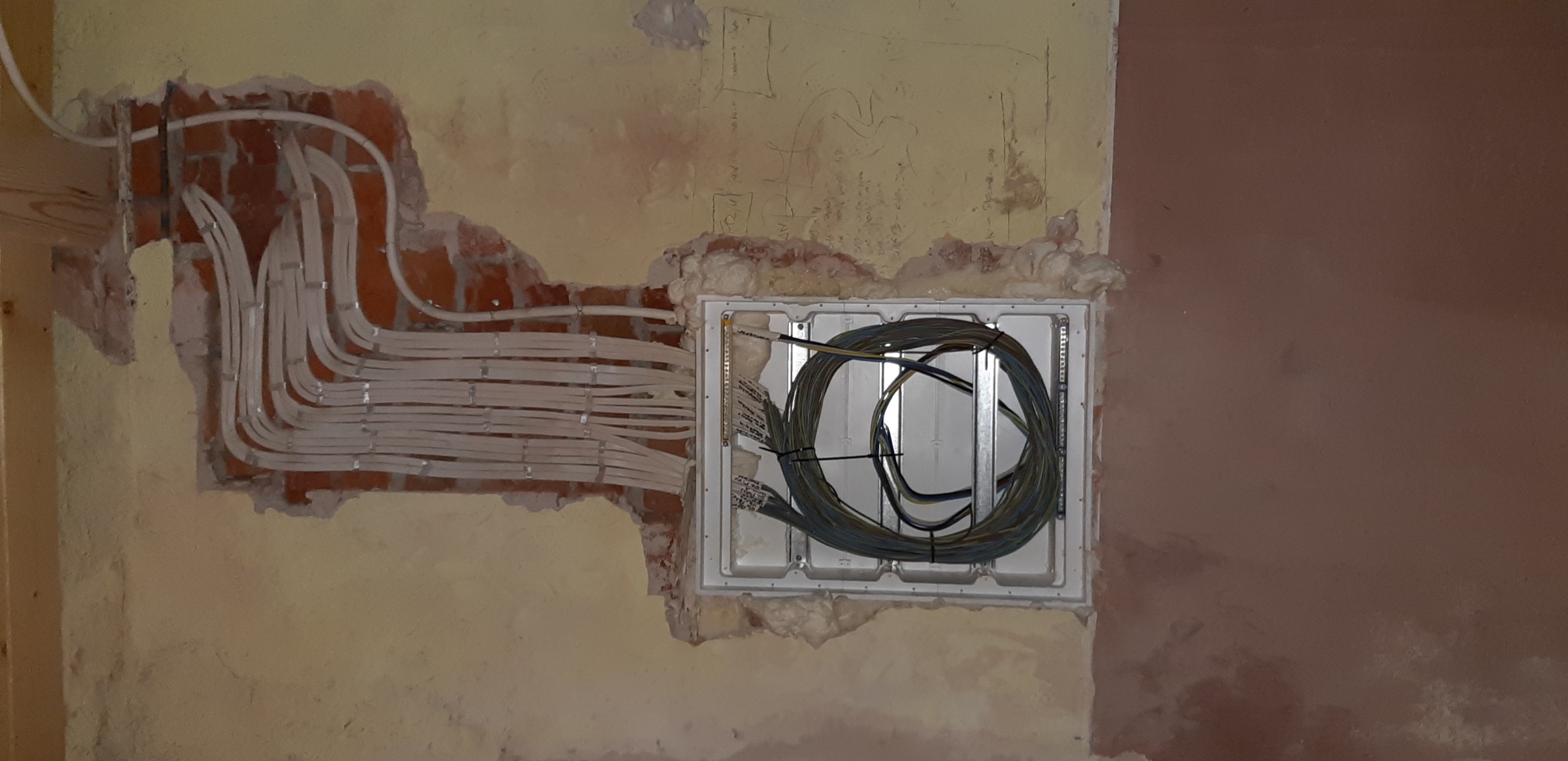 Wiązka białych kabli elektrycznych ułożonych w bruzdach w ceglanej ścianie, obok puszki instalacyjnej z nawiniętymi kablami, na tle surowych ścian w trakcie remontu.
