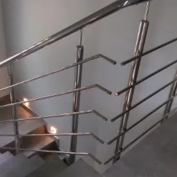 Nowoczesna balustrada schodowa ze stali nierdzewnej z poziomymi prętami zakończonymi pod kątem, zamontowana w jasnym wnętrzu z oknem i rośliną doniczkową.