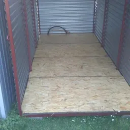 Wnętrze metalowego kontenera z podłogą z płyt OSB, widoczne z zewnątrz przez otwarty wjazd. Elementy konstrukcyjne pomalowane na czerwono, na zewnątrz rosnąca trawa.