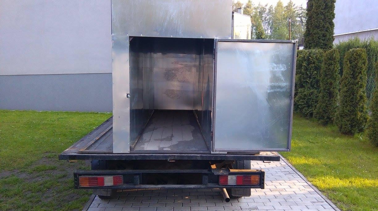 Metalowa zabudowa reklamowa na platformie samochodu dostawczego z otwartymi drzwiami, widok z tyłu na tle zieleni.