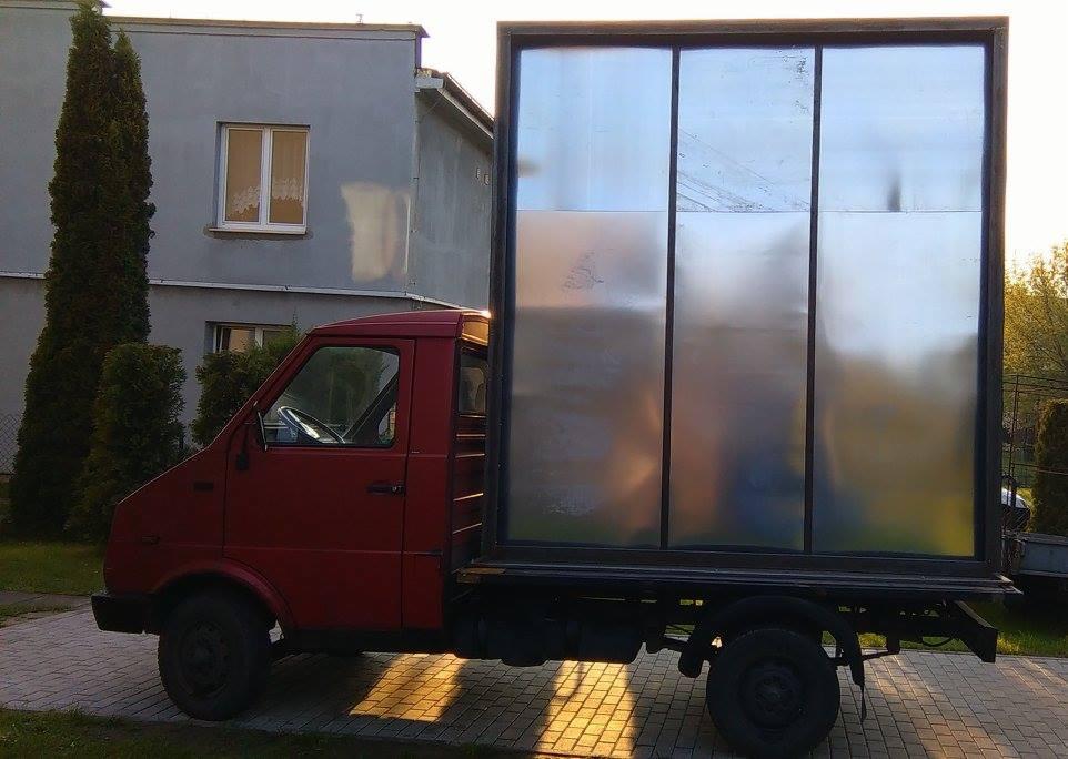 Czerwony samochód dostawczy z zabudową skrzyniową z blachy aluminiowej, zaparkowany na kostce brukowej przed budynkiem mieszkalnym.