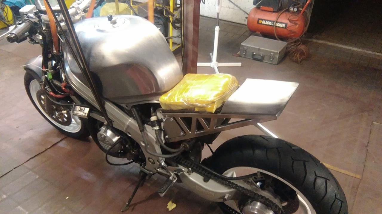 Motocykl w trakcie customizacji: surowy, metalowy zbiornik paliwa, spawana konstrukcja pod siedzenie, żółta folia ochronna na siedzeniu, widoczny kompresor w tle.