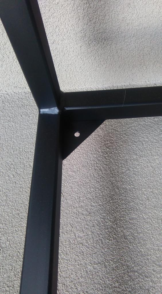 Czarny, metalowy stelaż pod umywalkę z otworem montażowym, spawany, na tle szarej, chropowatej ściany.