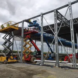 Stalowa konstrukcja hali w trakcie montażu, widoczne podnośniki koszowe z pracownikami, pochmurne niebo w tle.