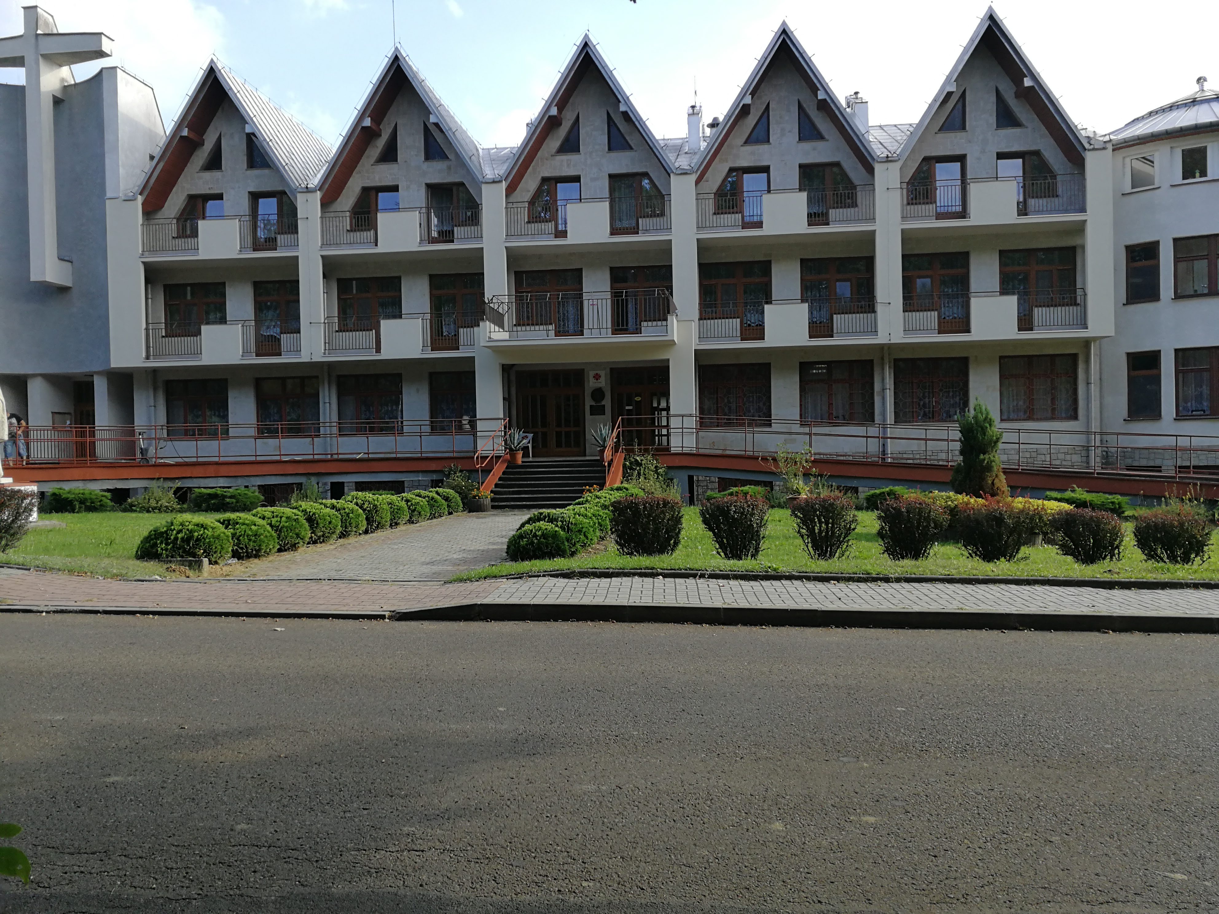 Trzypiętrowy budynek z powtarzalnymi segmentami z balkonami i trójkątnymi szczytami dachów, otoczony zielenią i chodnikiem, z widocznym fragmentem budynku kościelnego z krzyżem po lewej stronie.