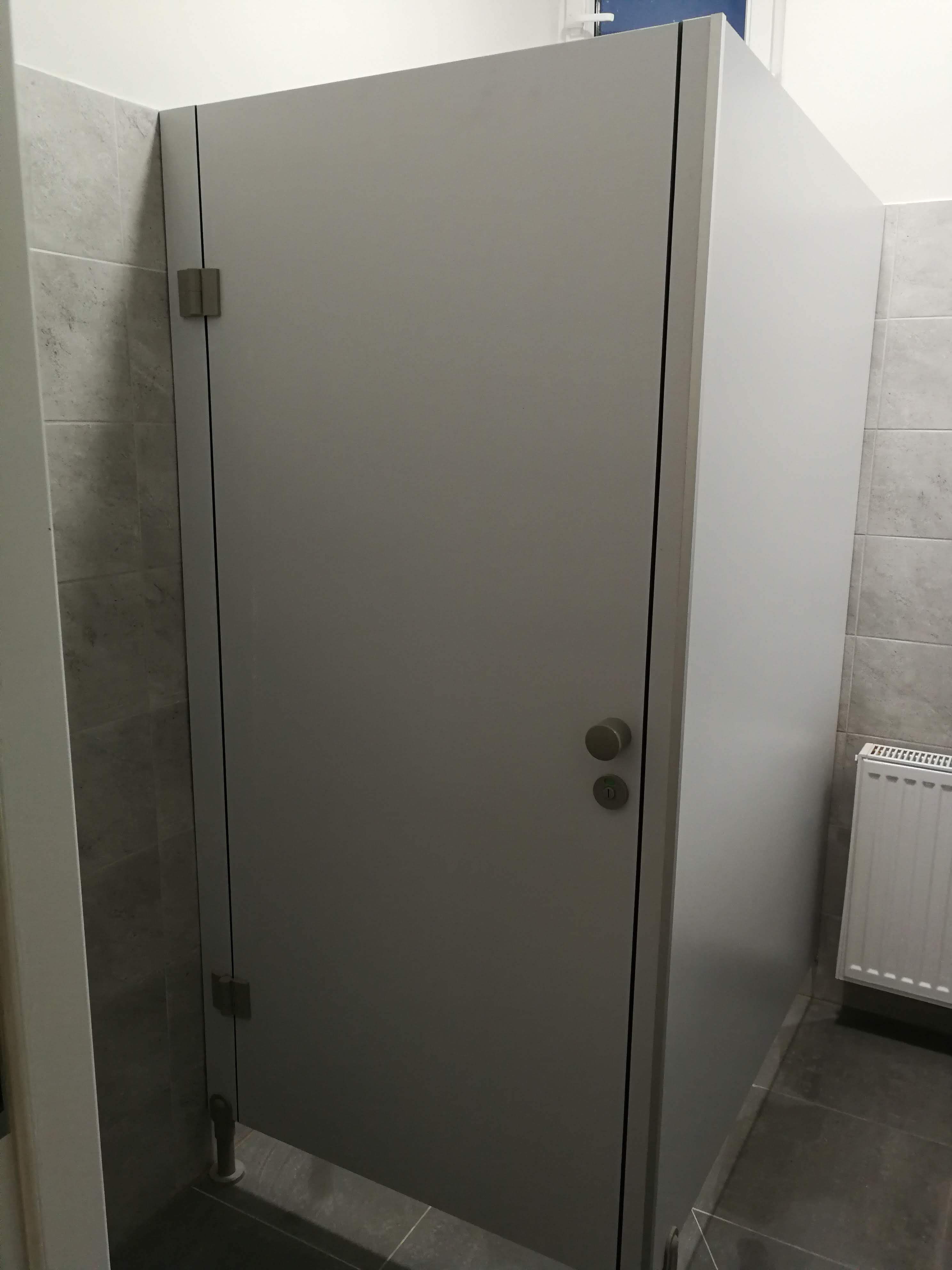 Szara kabina sanitarna z zamkiem i zawiasami w łazience wyłożonej szarymi płytkami, widoczny grzejnik.