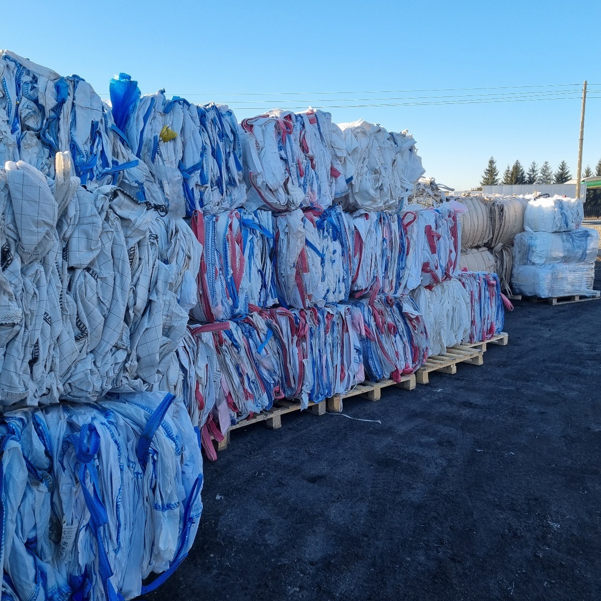 Ułożone na paletach, wypełnione białe worki typu big bag z niebieskimi i czerwonymi detalami, składowane na zewnątrz pod błękitnym niebem.