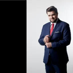 Mężczyzna z brodą, ubrany w granatowy garnitur i czerwoną koszulę w kropki, pozuje na jasnym tle, poprawiając mankiet marynarki.