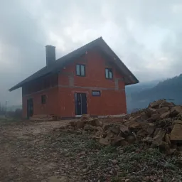 Dwukondygnacyjny dom w stanie surowym zamkniętym z czerwonej cegły, czarną dachówką i kominem, na tle pochmurnego nieba i zalesionych wzgórz, z hałdą kamieni na pierwszym planie.