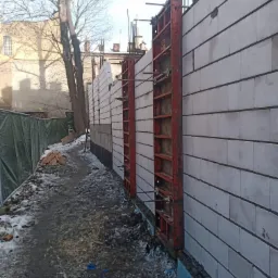 Wznoszenie ściany z bloczków betonowych z użyciem czerwonych szalunków, widoczna izolacja fundamentów i teren budowy pokryty resztkami śniegu.