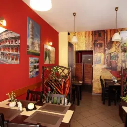 Wnętrze restauracji z czerwonymi ścianami zdobionymi wielkoformatowymi wydrukami Koloseum i Krzywej Wieży w Pizie, uzupełnione tapetą imitującą uliczkę włoskiego miasteczka.