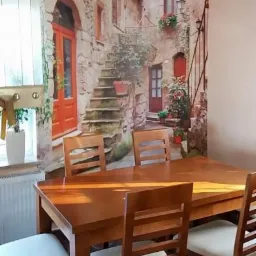 Jadalnia z fototapetą przedstawiającą włoską uliczkę z kamiennymi schodami i czerwonymi drzwiami, widoczny stół z krzesłami.