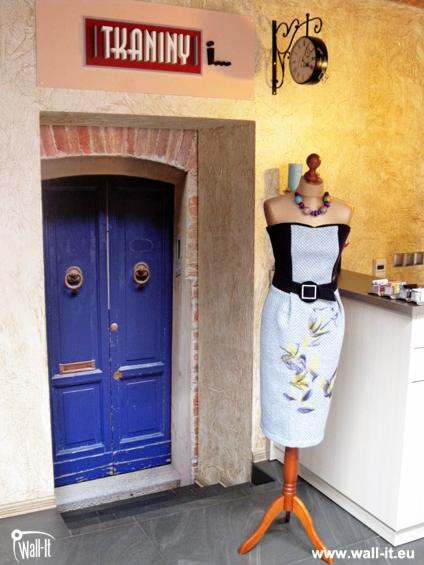 Fototapeta imitująca wejście do sklepu z tkaninami, z niebieskimi drzwiami i manekinem z sukienką w stylu vintage, zamontowana na ścianie.