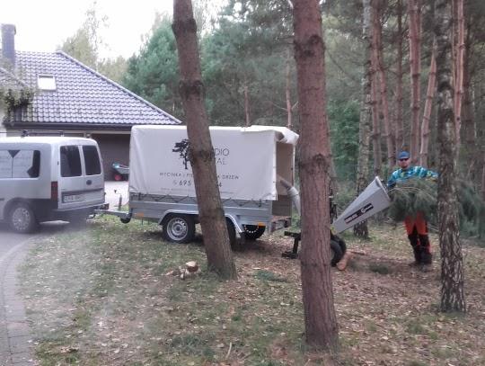 Pracownik w lesie wrzuca gałęzie do rębaka, obok przyczepa z plandeką i bus firmy.