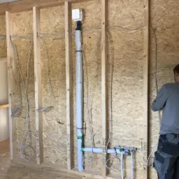 Instalacja przewodów elektrycznych i rur w ścianie szkieletowej budynku w trakcie budowy, widoczne zabezpieczenia i oznaczenia przewodów, pracownik w tle.