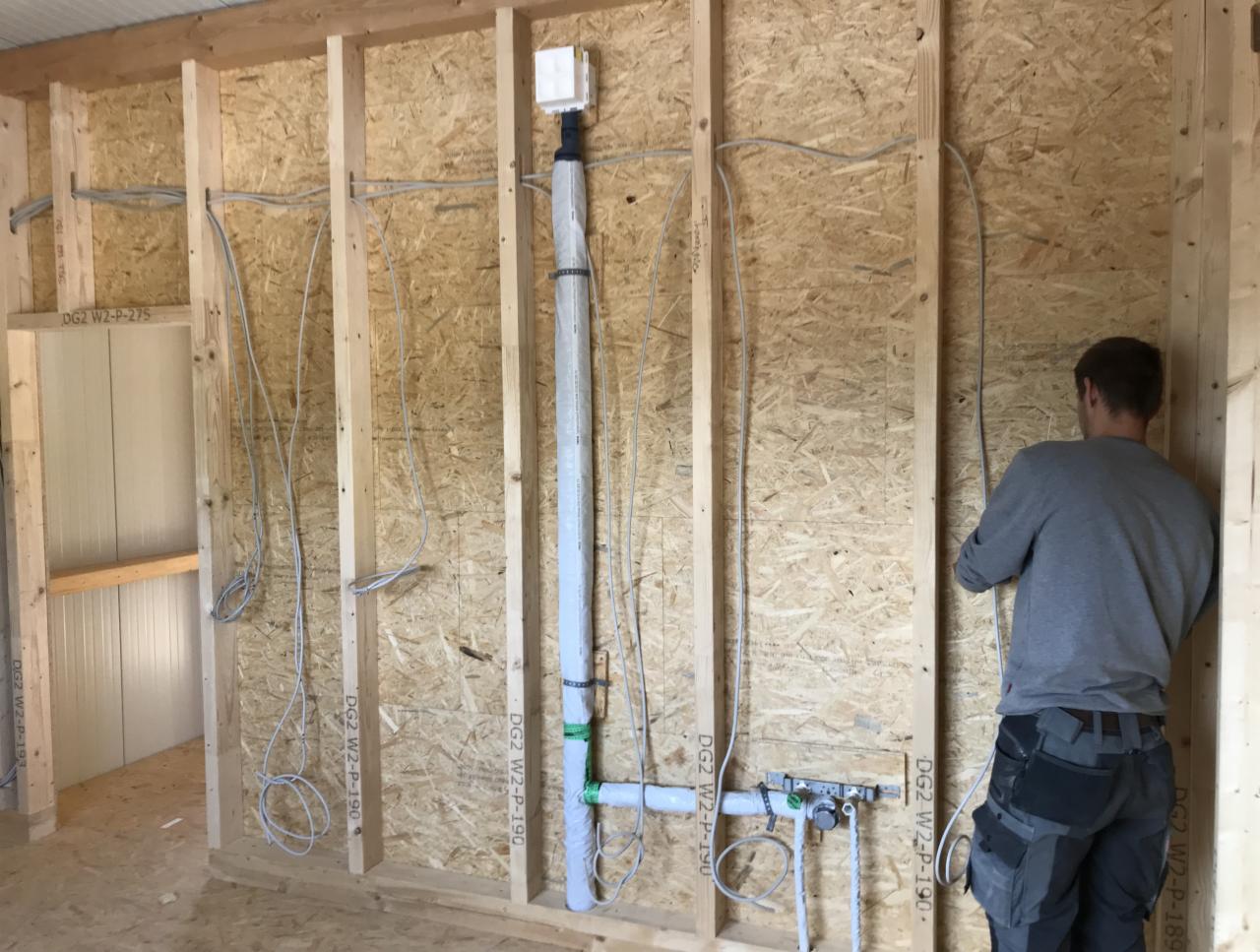 Instalacja przewodów elektrycznych i rur w ścianie szkieletowej budynku w trakcie budowy, widoczne zabezpieczenia i oznaczenia przewodów, pracownik w tle.