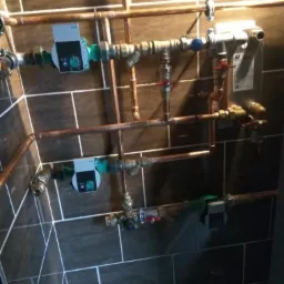 Kompleksowy system rur miedzianych z pompami obiegowymi i zaworami, zamontowany na ścianie z ciemnych, prostokątnych płytek imitujących kamień.