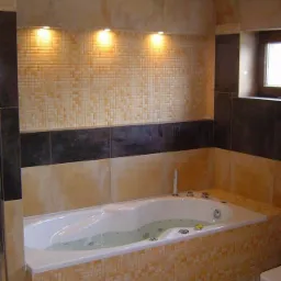 Wykończona łazienka z wanną z hydromasażem, mozaiką w odcieniach beżu i brązu, oświetleniem punktowym sufitowym i podłogowym, oraz wąskim oknem w ciemnej ramie.