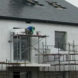 Ocieplanie elewacji budynku styropianem z rusztowania, widoczne okna i dach z dachówki.