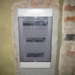 Zamontowana, nowa skrzynka rozdzielcza z widocznymi bezpiecznikami na ścianie z surową cegłą i resztkami tynku, poziomica częściowo widoczna w prawym dolnym rogu.