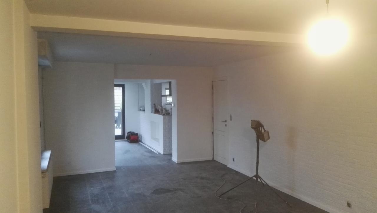 Wnętrze mieszkania po remoncie z pomalowanymi ścianami na biało, widoczny fragment kuchni w tle i lampa na statywie doświetlająca ścianę.