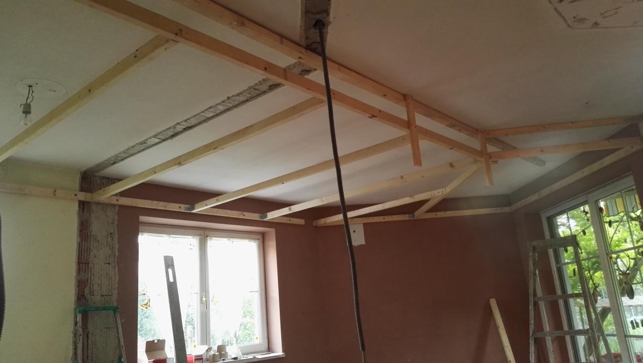 Wnętrze w trakcie prac przygotowawczych do montażu sufitu podwieszanego: widoczna drewniana konstrukcja szkieletowa przymocowana do sufitu i ścian, ściany częściowo otynkowane, okno z widokiem...