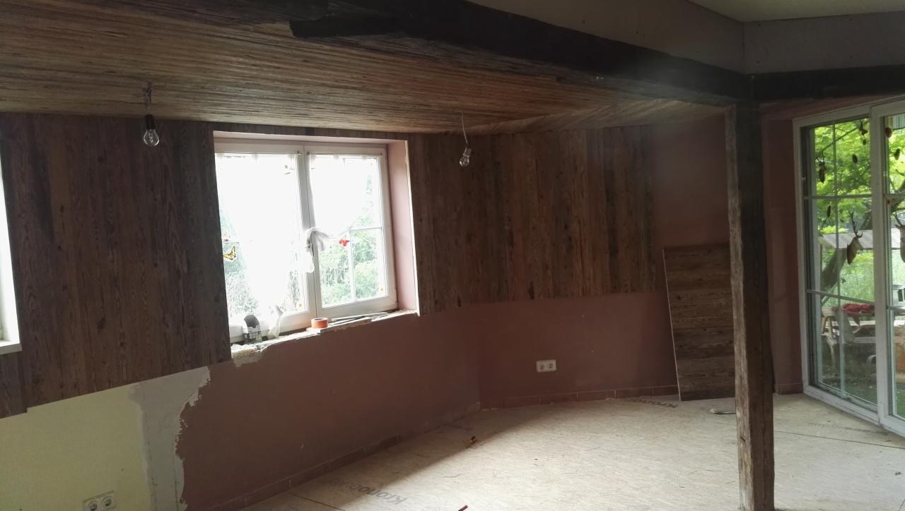 Wnętrze w trakcie remontu z drewnianą boazerią na ścianach i suficie, surową podłogą, oknem z widokiem na zieleń i wiszącą żarówką.