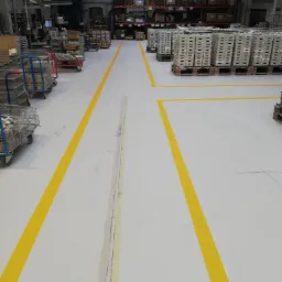 Wnętrze magazynu z białym podłożem i wyznaczonymi żółtymi pasami ruchu, w tle regały z kartonami i wózki transportowe