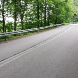 Asfaltowa droga z podwójną linią ciągłą, zabezpieczona metalową barierką, otoczona zielonymi drzewami w słoneczny dzień.