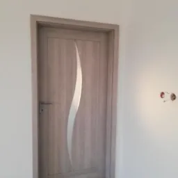 Zamknięte, jasne drzwi wewnętrzne z dekoracyjnym, pionowym, białym paskiem, zamontowane w świeżo wykończonym wnętrzu, widoczne przewody elektryczne w ścianie.