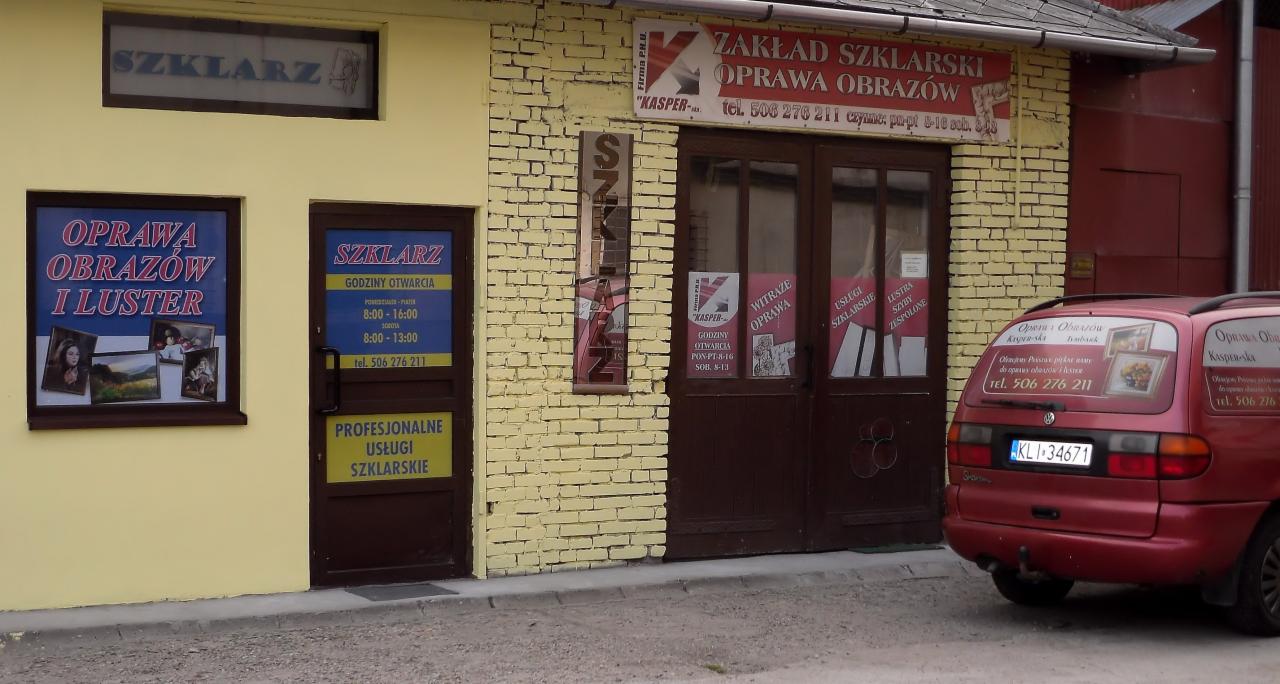 Fasada budynku z żółtej cegły z szyldami 'Zakład Szklarski Oprawa Obrazów' i 'Oprawa Obrazów i Luster', oklejony samochód z reklamą firmy szklarskiej na tle wejścia.