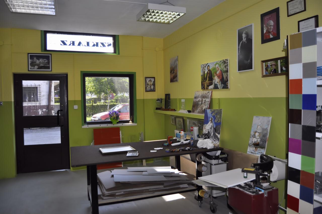 Warsztat z biurkiem, na którym leżą arkusze szkła lub pleksi, w tle okno z widokiem na samochód i kwiaty, na ścianach wiszą obrazy i zdjęcia.