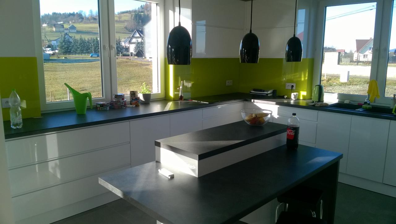 Nowoczesna kuchnia z białymi szafkami, ciemnym blatem i zielonym panelem szklanym, widok na wiejski krajobraz za oknem, czarne lampy wiszące nad wyspą kuchenną.