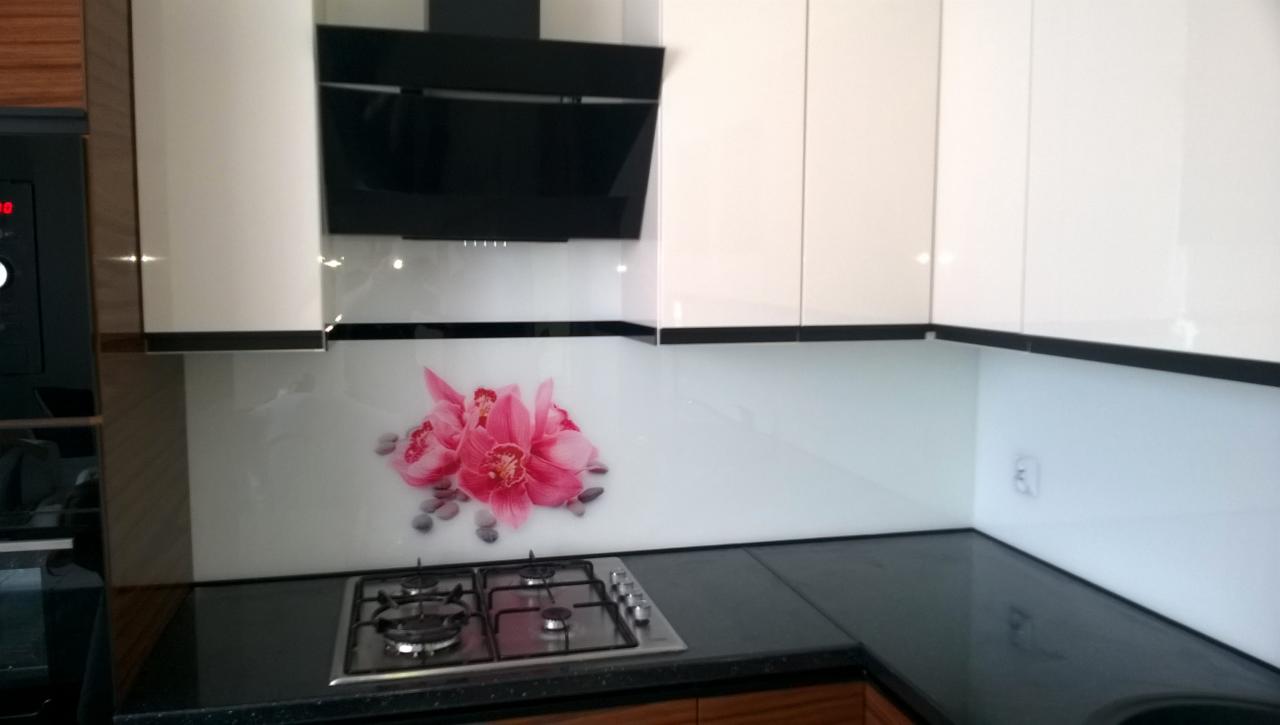 Szklany panel kuchenny z motywem różowych kwiatów orchidei, zamontowany nad czarnym blatem i kuchenką gazową, w nowoczesnej kuchni z białymi szafkami i czarnym okapem.