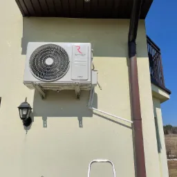 Biała jednostka zewnętrzna klimatyzacji Rotenso Versu Series zamontowana na elewacji domu, widoczna rynna, lampa zewnętrzna i fragment drabiny.