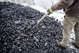 Mężczyzna łopatą nabiera węgiel z pryzmy na tle śniegu. Widoczne fragmenty ubrania roboczego i rękawice ochronne.