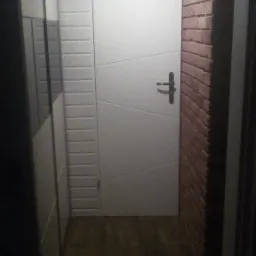 Wąskie wnętrze z białą, geometryczną ścianą i drzwiami, kontrastującą ceglaną ścianą oraz podłogą imitującą drewno.